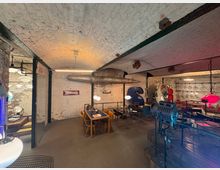 Industrie-inspiriertes Interieur in einem Raum mit gewölbter Ziegeldecke, sichtbaren Rohren und Wänden aus rohem Ziegelmauerwerk. Die Einrichtung umfasst moderne und retro designte Möbel, bunte Dekorationen und Lampen in einem offenen, kreativ gestalteten Bereich.