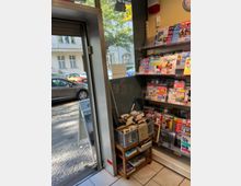 Ein kleines Geschäft mit einem Zeitungs- und Zeitschriftenregal rechts im Bild, gefüllt mit bunten Titeln. Links im Bild ist eine offene Tür, die auf eine Straße mit geparkten Autos und Fußweg mit Herbstlaub blickt. Vor dem Regal steht ein Regal mit Büchern und einem Schild mit der Aufschrift „Jedes Teil 1 €“.