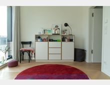 Heller Wohnraum mit einem weißen Sideboard, das mit Büchern, Dekorationsgegenständen und gerahmten Bildern bestückt ist. Links steht ein schwarzer Stuhl mit einer Decke, daneben eine große Fensterfront mit Vorhängen; der Boden ist mit hellem Holzparkett und einem roten Teppich ausgestattet.