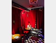 Ein kleiner Innenraum mit roten Vorhängen an den Wänden, schwarz-weiß kariertem Boden und roten Stühlen an runden Holztischen. Im Hintergrund ist eine skulpturartige Büste mit Lampe auf einem Tisch zu sehen.