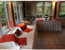 Ein heller Raum mit großen Fenstern, dekoriert für ein Buffet. Weiße Tische mit orangenen Läufern und silbernen Warmhaltebehältern sind aufgestellt, ergänzt durch ein Kerzenständer und Blumengesteck im Vordergrund.