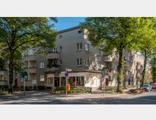 Eckgebäude in einer Wohnstraße mit beigefarbener Fassade, Balkonen und einem Laden im Erdgeschoss. Vor dem Gebäude stehen Bäume, die Schatten spenden, und es gibt eine Verkehrsampel sowie parkende Autos auf der Straße.