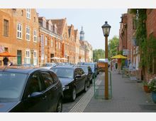 Straßenszene in einer historischen Altstadt mit roten Backsteingebäuden auf beiden Seiten, zahlreichen geparkten Autos und Gehwegen mit Schildern, Geschäften und Sonnenschirmen. Im Hintergrund sind Bäume und ein markanter Turm sichtbar.