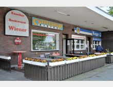 Backsteingebäude mit mehreren Schildern, darunter "Coffeeshop Bistro" und "Kogge", sowie Angeboten wie Bockwurst, Bouletten und Salaten. Vor dem Gebäude befinden sich Holzbänke und Blumenkästen, die mit gelben und orangefarbenen Blumen bepflanzt sind.