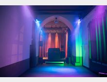 Innenraum eines Veranstaltungssaals mit einer Bühne, eingerahmt von einem großen Bogen. Die Bühne ist mit einem Vorhang, Lautsprechern und einer Lichtanlage ausgestattet, während bunte Lichter und Nebel den Raum beleuchten.