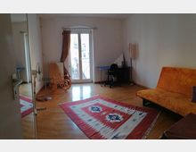 Helles Wohnzimmer mit Holzboden, einem roten Teppich mit geometrischem Muster, einer orangenen Couch, einem Schaukelstuhl und einem kleinen Schreibtisch mit Stuhl in der Ecke. Eine Doppeltür mit Glaseinsatz führt zu einem schmalen Balkon.