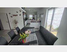 Moderne Wohnküche mit Esstisch und Eckbank im Vordergrund, darauf eine Vase mit gelben Tulpen. Im Hintergrund eine weiße Einbauküche mit Fenstern, links eine Zimmertür und rechts eine Terrassentür mit halbtransparenten Vorhängen.