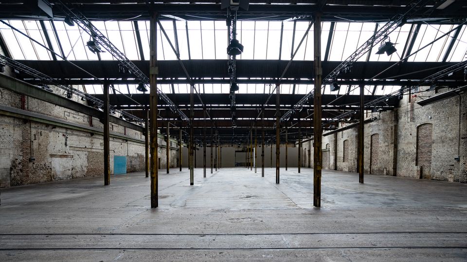Large empty industrial warehouse interior with a concrete floor, exposed brick walls, and rows of metal support columns. A high roof with skylights and suspended lighting trusses spans the open space.