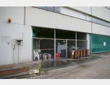 Industriegebäude mit einem offenen Unterstand, der mit Maschendrahtzaun abgesichert ist. Im Vordergrund stehen mehrere braune Mülltonnen, daneben sind gestapelte weiße Plastikstühle, Metallfässer und andere gelagerte Gegenstände zu sehen; der Boden ist teils gepflastert und teils betoniert.