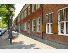 Reihe historischer Backsteinhäuser mit weißen Fenstern und vereinzelt hölzernen Fensterläden, entlang einer gepflasterten Straße mit Bäumen und geparkten Autos. Im Vordergrund steht eine schwarze Laterne.