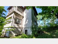 Zweistöckiges Wohnhaus mit weißem Putz und einem Abschnitt aus Holzfassade auf der linken Seite, umgeben von dichtem Laubwerk und Bäumen. Im Vordergrund sind Fenster mit Holzrahmen und ein kleiner, ungenutzter Gartenbereich sichtbar.