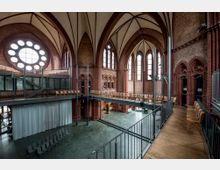 Innenraum einer historischen Kirche mit neugotischer Architektur, rotem Ziegelmauerwerk und Spitzbögen. Der Raum ist mit modernen Elementen wie einem Zwischengeschoss aus Stahl und Holzstühlen ausgestattet, und große Rund- und Spitzbogenfenster lassen viel Licht herein.
