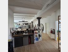 Großer, offener Raum mit hell gestrichenen Wänden und freiliegender weißer Decke mit gewölbten Balken. Das Interieur umfasst eine Küchenzeile mit Spüle und Geräten, ein Regal mit Ablagen, Holzboden und einzelne Säulen, die den Raum strukturieren; im Hintergrund befinden sich Arbeitsbereiche mit Schreibtischen.