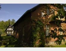 Backsteinbau mit geschmücktem Ziegelmuster und begrüntem Efeu an der Fassade, daneben ein schwarzes Fachwerkhaus mit weißen Fensterrahmen, eingebettet in eine grüne, von Bäumen umgebene Landschaft. Im Vordergrund ein gepflegter Rasen mit einzelnen Sträuchern.