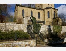 Gelbsteinige Kirche mit hohen Bogenfenstern, umgeben von einer Natursteinmauer, teilweise bewachsen mit Efeu. Ein geschmiedetes Metalltor und eine Treppe führen durch die Mauer zum erhöhten Eingangsbereich, umgeben von gepflegten Sträuchern.