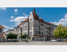 Historisches Wohn- und Geschäftsgebäude mit Zierelementen und roten Ziegeldächern an einer Straßenecke; im Vordergrund ein Kopfsteinpflasterplatz mit Bäumen, Lampen und geparkten Autos. Der Himmel ist klar mit vereinzelten Wolken.