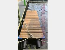 Ein schmaler, rechteckiger Holzsteg schwimmt auf dem Wasser und wird von Seilen befestigt. Der Steg hat blaue Polster an den Ecken sowie Metallhalterungen, und im Hintergrund sind weitere Stege und das Wasser zu sehen.