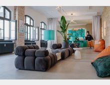 Heller Lounge- und Barbereich in einer umgebauten Industriehalle mit hohen Rundbogenfenstern, freigelegten Ziegelwänden und Betonboden. Große modulare Sofaelemente, türkisfarbene Leuchten, eine große Topfpflanze und eine türkisfarbene Bar mit beleuchteten Regalen prägen den Raum.