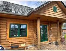 Holzhaus mit Blockbohlenfassade, grün gerahmten Fenstern und einer grünen Eingangstür. Der überdachte Eingangsbereich wird von massiven Holzsäulen getragen, daneben stehen Bänke, Fahrräder und dekorierte Gegenstände.