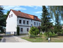 Zweistöckiges weißes Gebäude mit rotem Satteldach und mehreren Fenstern, umgeben von Bäumen und kleinen Sträuchern. Im Vordergrund führt ein Weg mit einem Tor zum Gebäude, rechts steht eine Rasenfläche mit vereinzelten Pflanzen.