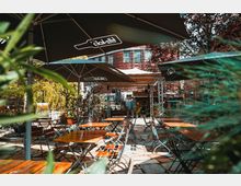 Außenbereich eines Cafés mit mehreren Holztischen und klappbaren Stühlen auf einem gepflasterten Boden. Große Sonnenschirme spenden Schatten, im Hintergrund sind Backsteingebäude, Pflanzen und eine überdachte Bühne zu sehen.