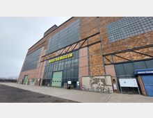 Industriegebäude aus Ziegelsteinen mit großen, rechteckigen Fensterflächen und der Aufschrift "Industriemuseum" in gelben Buchstaben über dem Eingang. Der Vorplatz ist gepflastert, und eine moderne Gebäudeverkleidung ergänzt das historische Erscheinungsbild.