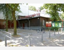 Außenansicht einer Gaststätte mit hölzerner Fassade, rotem Schriftzug „Gaststätte“ auf dem Dach und einem Fahrradständer im Vordergrund. Links und rechts der Eingangstür befinden sich Schilder mit Öffnungszeiten und Informationen; umgeben von Bäumen und Metallgeländern.