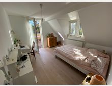 Helles Schlafzimmer mit Dachschrägen, einem Bett mit rosa Bettwäsche, einem Schreibtisch und einer Kommode. Eine geöffnete Balkontür führt zu einem kleinen Balkon mit Tisch und Stühlen; Fenster bieten Ausblick auf Bäume.