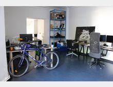 Büro mit mehreren Arbeitsplätzen, blauer Regalwand und einem Fahrrad im Vordergrund. Die Einrichtung umfasst Schreibtische mit Computern, Bürostühle und unterschiedliche Gegenstände auf den Tischen sowie an der Wand.