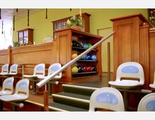 Bowlingbahn mit Sitzplätzen aus weißem Kunststoff und blauen Rückenlehnen, einer Holztheke im Hintergrund sowie Regalen für bunte Bowlingkugeln. Ein Geländer und einige Stufen führen zur Erhöhung mit der Theke.