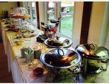 Ein Buffet mit verschiedenen Speisen, serviert auf einer langen, mit einem Tischtuch bedeckten Tafel. Im Vordergrund stehen glänzende Warmhaltebehälter, im Hintergrund sind Teller, Schalen, und unter Abdeckhauben präsentierte Speisen sowie Fenster mit Blick ins Grüne zu sehen.