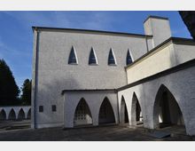 Moderne Kirche mit weißer, rau verputzter Fassade, spitzbogigen Fenstern und Arkaden mit Spitzbögen. Der Vorplatz ist gepflastert und von Bäumen umgeben.