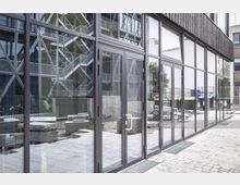 Moderne Glasfassade eines Gebäudes mit bodentiefen Fenstern und Türen. Im Hintergrund sind ein Außentreppenhaus mit Metallstruktur und Baustellenelemente wie Betonblöcke und Müllbehälter zu erkennen.