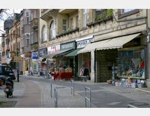 Straßenszene mit einer Reihe von Altbauten, in deren Erdgeschossen verschiedene Geschäfte liegen, darunter ein Antiquariat und ein Laden mit Auslagen vor der Tür. Der Gehweg ist mit Pflastersteinen gestaltet, teils überdacht durch Markisen, und es sind geparkte Fahrzeuge sowie Fahrradbügel zu sehen.