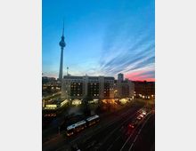 Abendansicht einer städtischen Straße mit beleuchteten Gebäuden, darunter ein mehrstöckiges Wohn- und Geschäftshaus. Im Hintergrund ist der Berliner Fernsehturm sichtbar, während der Himmel in Blau- und Rottönen gefärbt ist.