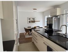 Modern open-plan kitchen and dining area with white cabinetry, a black countertop island with sink and faucet, and stainless steel appliances. In the background, a dining table with chairs sits under a pendant light near a window, with a framed picture on the wall and wood flooring.