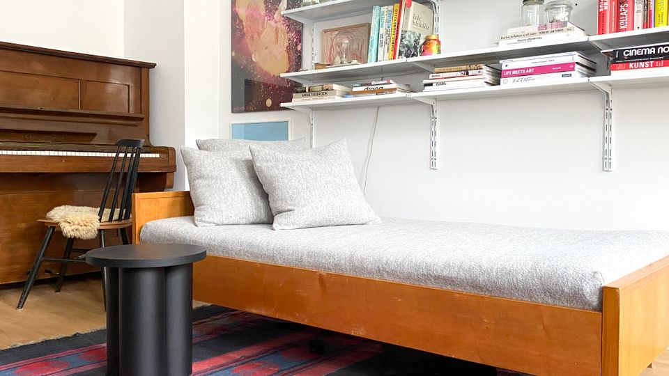 Helles Wohnzimmer mit Holz-Recamiere und grauen Kissen auf gemustertem Teppich, daneben ein schwarzer Beistelltisch. An der Wand befinden sich offene Regale mit Büchern und Gläsern; links steht ein Klavier neben einem Stuhl.