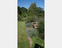 Ein gepflegter Garten mit einer Holzbank, umgeben von Blumenbeeten mit lila Blüten und grünen Sträuchern. Im Hintergrund sind größere Bäume und ein klarer blauer Himmel zu sehen.