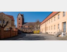 Straßenszene mit einer historischen Backsteinkirche im Zentrum, flankiert von einem gelben Wohngebäude und einem langen, hellen Verwaltungsgebäude. Neben der gepflasterten Straße befinden sich kahle Bäume und ein niedriger Backsteinzaun.