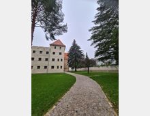 Gepflasterter Weg in einer gepflegten Grünanlage, flankiert von Rasenflächen und Bäumen, mit einem historischen, quadratischen Gebäude aus hellem Putz und roten Dachziegeln im Hintergrund. Im hinteren Bereich ist eine hohe Mauer sichtbar.