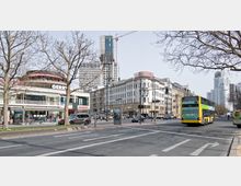 Straßenszene in einer Stadt mit mehrspuriger Fahrbahn, links Geschäfte mit großen Fenstern und Aufschriften, darunter "Gerry Weber", rechts ein mehrstöckiges Hotelgebäude. Im Hintergrund sind moderne Hochhäuser mit Kranauslegern sichtbar, im Vordergrund fahren ein gelber Doppeldeckerbus und mehrere Autos, während kahle Bäume die Straße säumen.