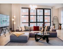 Modernes Wohnzimmer mit hellem Holzboden, einer grauen Eckcouch mit bunten Kissen, einem schwarzen Couchtisch mit Glasplatte und einer Stehlampe mit Kugelleuchten. Im Hintergrund befinden sich große Fenster mit Blick auf ein benachbartes Gebäudefassaden und ein Standspiegel neben Dekovasen.