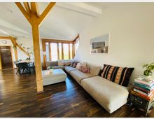 Wohnzimmer in einem Dachgeschoss mit sichtbaren Holzbalken, dunklem Holzboden und großen Fenstern, die viel Tageslicht hereinlassen. Eine große beigefarbene Couch mit bunten Kissen steht gegenüber einem kleinen Stapel Bücher mit einer Pflanze darauf. Im Hintergrund ein Esstisch mit schwarzen Stühlen und Pflanzen an der Wand.