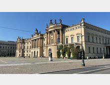 Neoklassizistisches Gebäude mit Säulenfassade und zahlreichen Statuen auf dem Dach, umgeben von einem gepflasterten Platz bei klarem Himmel; im Vordergrund ist eine Straßenlaterne zu sehen.