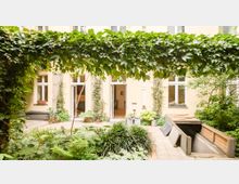 Ein begründer Innenhof mit Kletterpflanzen, die an einer Pergola entlangranken, und einer zentralen Fläche mit Farnen und anderen Pflanzen. Im Hintergrund befindet sich eine beige Fassade mit großen Fenstern und offenen Türen, die in ein Gebäude führen.