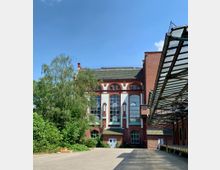 Industriegebäude aus rotem Backstein mit großen, hohen Fenstern und verzierten weißen Elementen; im Vordergrund ein gepflasterter Hof mit angrenzender Überdachung aus Glas und Metall sowie grüne Bepflanzung.