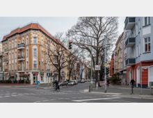 Straßenszene mit einem Eckhaus im historischen Stil, gelb verklinkert mit weißen Ornamenten und roten Dachziegeln. Umgeben von weiteren mehrstöckigen Wohngebäuden und kahlen Bäumen, Kreuzung mit Ampeln, Fahrradfahrer und Fußgänger, Straßenschild mit "Wollankstraße".