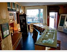 Eine kleine, gemütliche Bar mit rustikalem Interieur, darunter eine abgenutzte Theke mit Gläsern, Tafeln mit Speise- und Getränkemenüs an den Wänden und große Fenster mit Blick nach draußen. Die warme Beleuchtung und Holzelemente bestimmen das Ambiente.