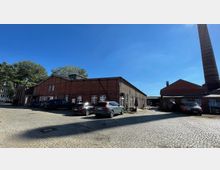 Backsteingebäude einer alten Industrieanlage mit schrägem Satteldach und weißen Fenstern, umgeben von gepflastertem Hof, auf dem einige Autos geparkt sind; im Hintergrund sind Bäume und ein hoher Schornstein zu sehen.