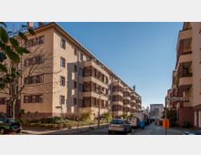 Wohnblock im Stil der klassischen Moderne mit beigefarbenen Putzfassaden, rotbraunen Klinkerelementen und Balkonen. Die Straße ist von geparkten Autos gesäumt, im Vordergrund stehen Straßenschilder und ein Baum mit kahlen Ästen.